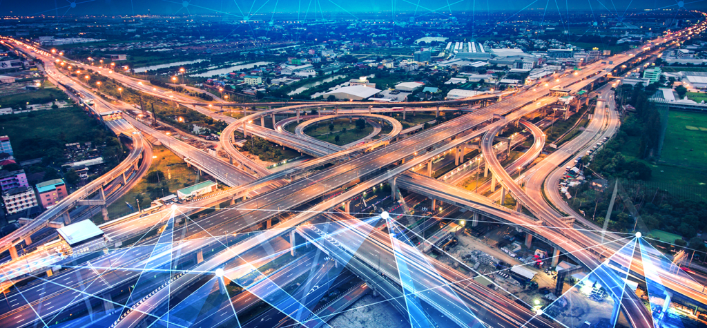 Aerial view of a large, complex motorway interchange with multiple curving slip roads and flyovers lit by streetlights at dusk. Overlay of digital blue lines and nodes suggests connectivity, technology, or smart city infrastructure integration.