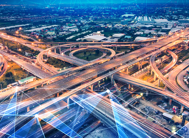 Aerial view of a large, complex highway interchange with looping overpasses and multiple lanes, surrounded by buildings and greenery at dusk. Digital blue lines and nodes from Spectrum Roadmap Studio are superimposed, suggesting technology or connectivity.