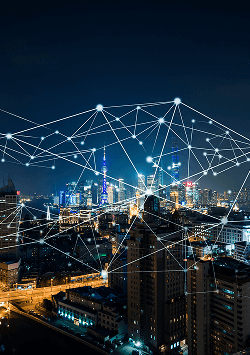 A nighttime cityscape with illuminated skyscrapers and streets. White glowing dots and connecting lines are superimposed over the scene, symbolizing a digital network and connectivity across the urban environment.