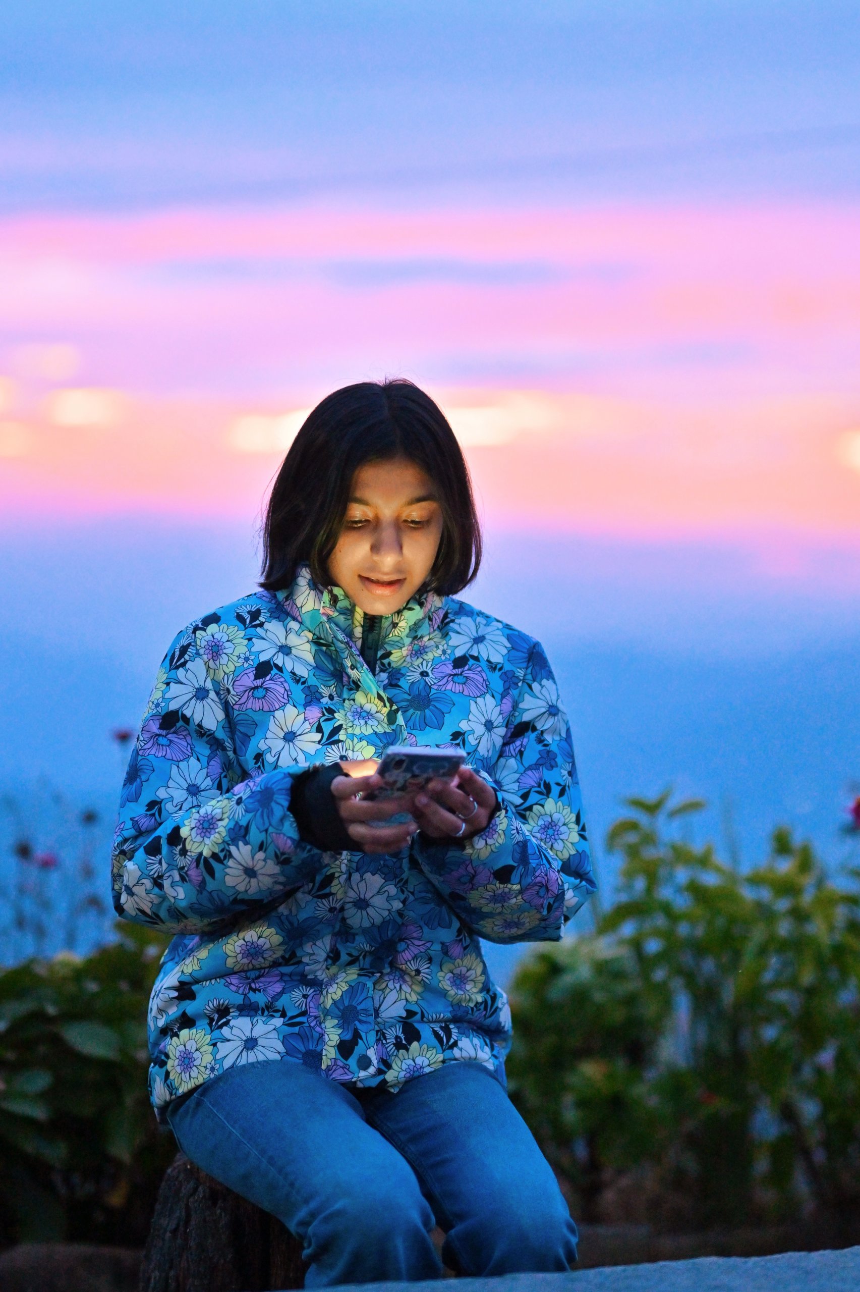 Young girl in a floral jacket looks at her mobile, possibly exploring the latest updates on GSMA Spectrum, glowing at dusk with a colourful sunset in the background.