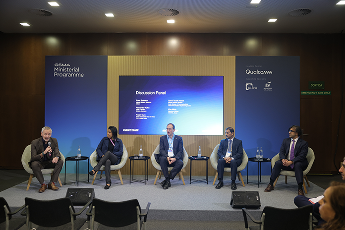 Five panellists sit on stage with microphones during a discussion at a conference, screen behind them.