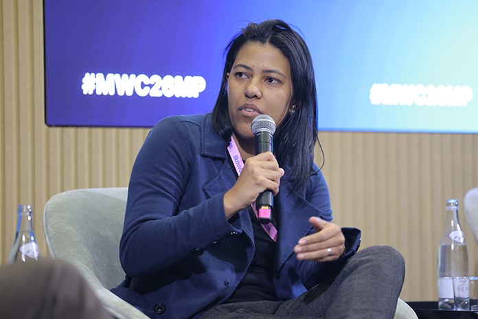 Person sitting and speaking into a microphone on a panel, with #HWC20AMP on a screen in the background.