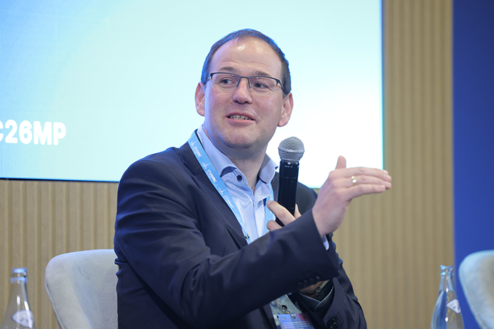 A man in a suit speaks into a microphone whilst gesturing with his hand during a panel discussion.