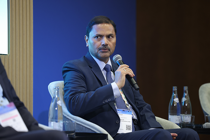 Man in a suit speaking into a microphone at a panel discussion, seated beside water bottles.