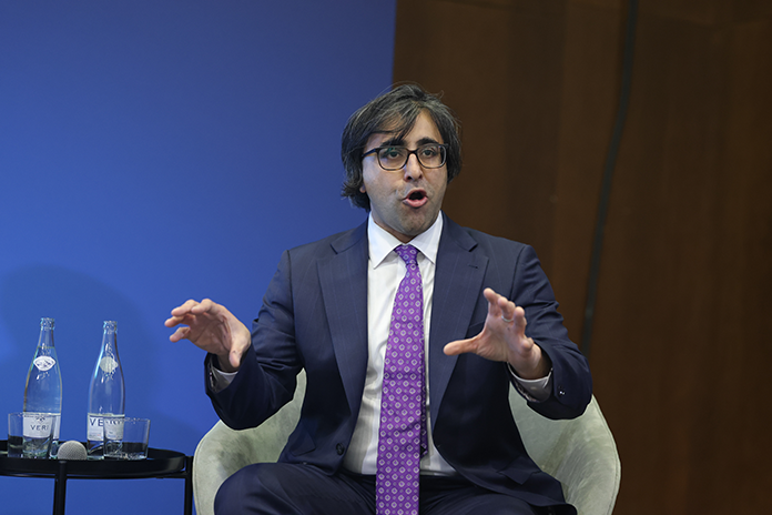Man in a suit speaks animatedly while seated, with water bottles on a table beside him and a blue background.