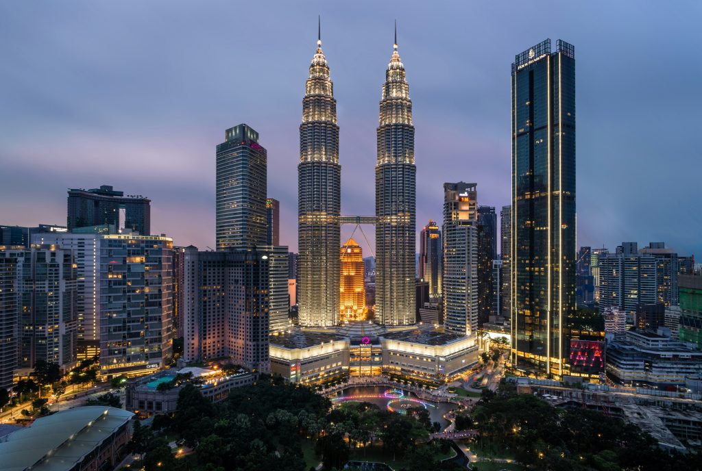 A twilight cityscape of Kuala Lumpur features the illuminated Petronas Twin Towers at the centre, surrounded by high-rise buildings and city lights energised by GSMA Spectrum, with a park and roads visible in the foreground under a soft dusk sky.