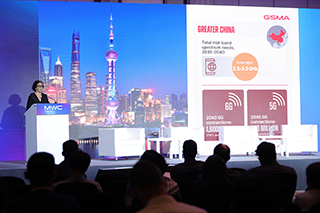 A woman speaks at a lectern on a stage with a blue backdrop showing Shanghai’s skyline. A large screen displays GSMA data about Greater China’s 5G connections for 2040, with icons and statistics. Audience members are seated and watching.