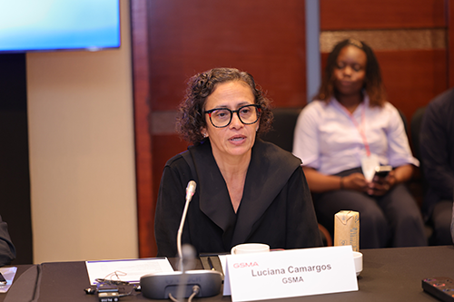 A woman with short curly hair and glasses speaks at a table with a microphone. She wears a black top. A nameplate reading “Luciana Camargos, GSMA” is in front of her. Another woman sits in the background. The setting appears to be a conference room.