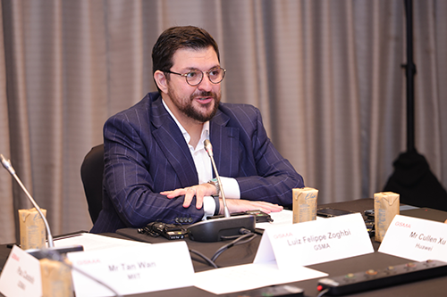 A man with glasses and a trimmed beard, dressed in a blue pinstripe suit, sits at a conference table speaking. Nameplates, microphones, and drink cartons are visible on the table; a beige curtain forms the background.