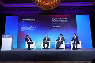 Four men in business attire sit on a stage panel with blue lighting and a chandelier overhead. A lectern labelled “MWC” is on the left. Names and titles of the speakers are displayed on the screen behind them. The setting is formal and professional.