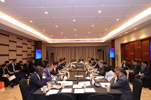 A large group of people in formal attire sit around a U-shaped conference table in a well-lit room, engaged in discussion. Laptops, documents, and drinks are on the tables. Two screens display content at the front. Curtains cover the back wall.