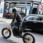A person wearing a black helmet and clothing rides a black motorcycle on a city street. In the background, there's a café named "Bloom" and a speed limit sign displaying "20." A black SUV is nearby, and pedestrians are seated outside the café.