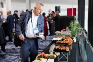 A man in a blue suit with a bag over his shoulder holds a plate and a drink while selecting food from a buffet table. The table has pastries, fruit, and small desserts. Several people in business attire mingle in the background of a well-lit, modern room.