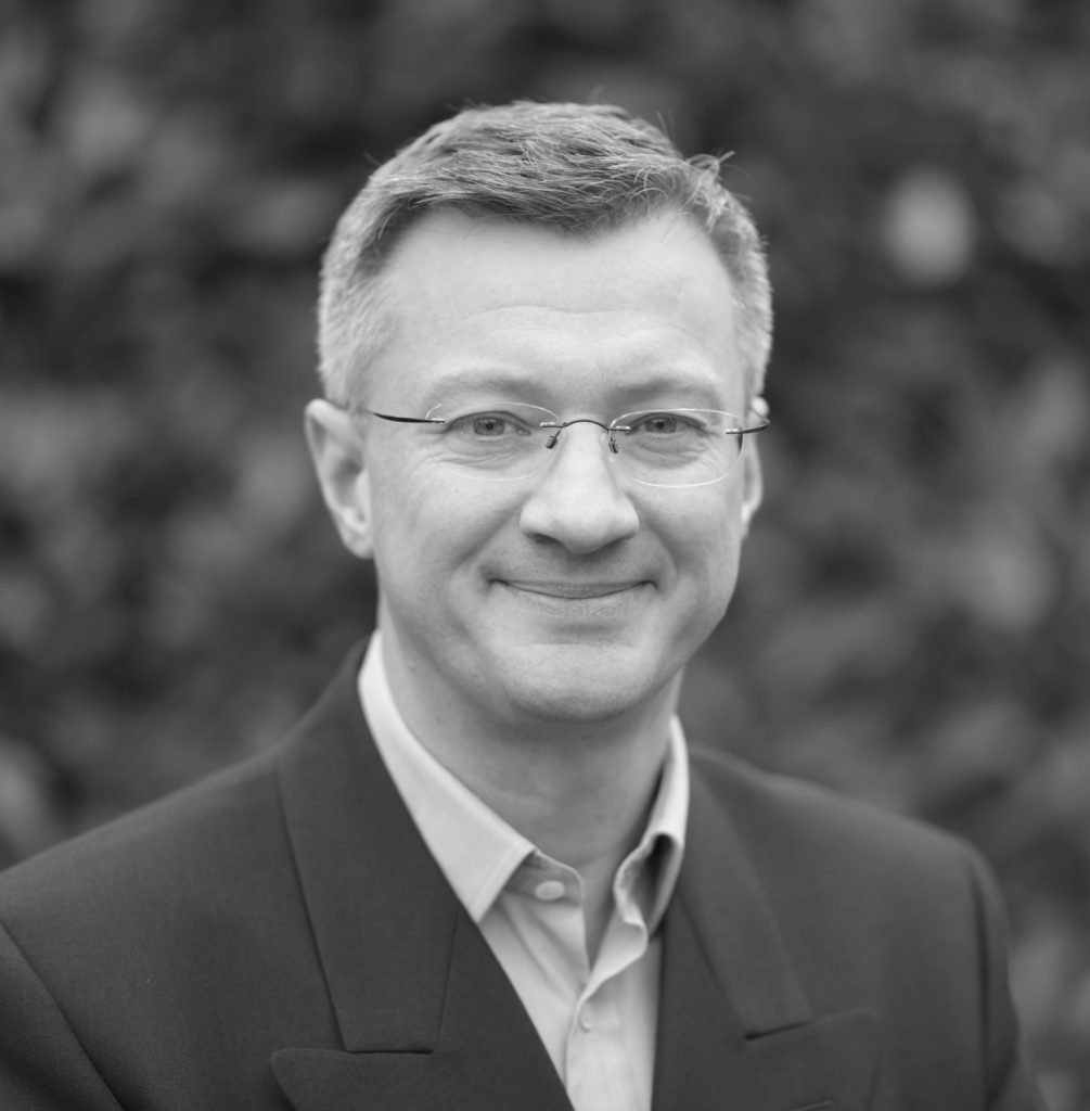 A grayscale portrait of a person with short hair and glasses, wearing a suit jacket over a light-colored shirt. They are smiling gently, and the background is softly blurred, enhancing focus on the subject's face and attire.