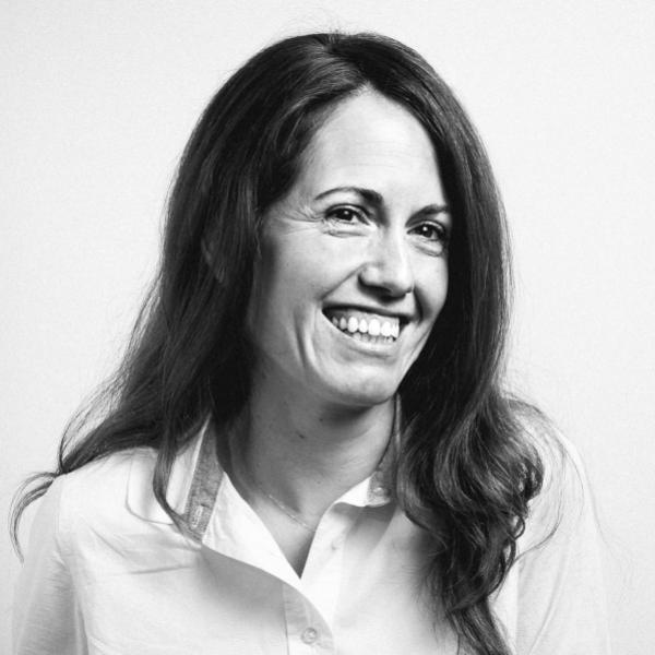 Black and white portrait of a woman smiling. She has long, straight hair and is wearing a light-colored button-up shirt. The background is plain, emphasizing her bright expression and relaxed pose.