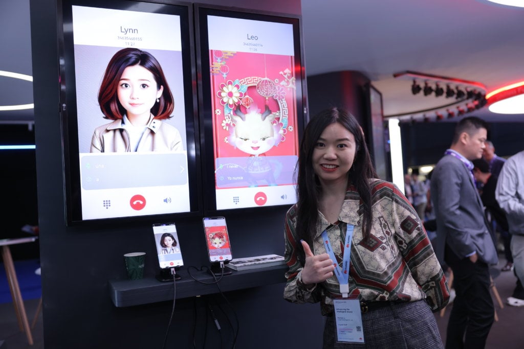 A woman stands smiling and giving a thumbs-up beside two large screens displaying cartoon avatars labeled "Lynn" and "Leo" with video call interfaces. Below, two smartphones connected to the screens mirror similar images. The setting appears tech-centric, with people in the background.