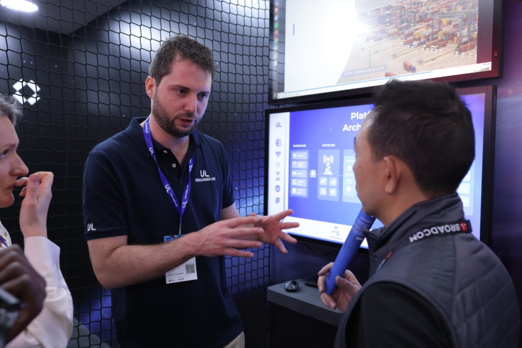 A person gesturing while explaining a point to another individual at a technology showcase with a digital display in the background.