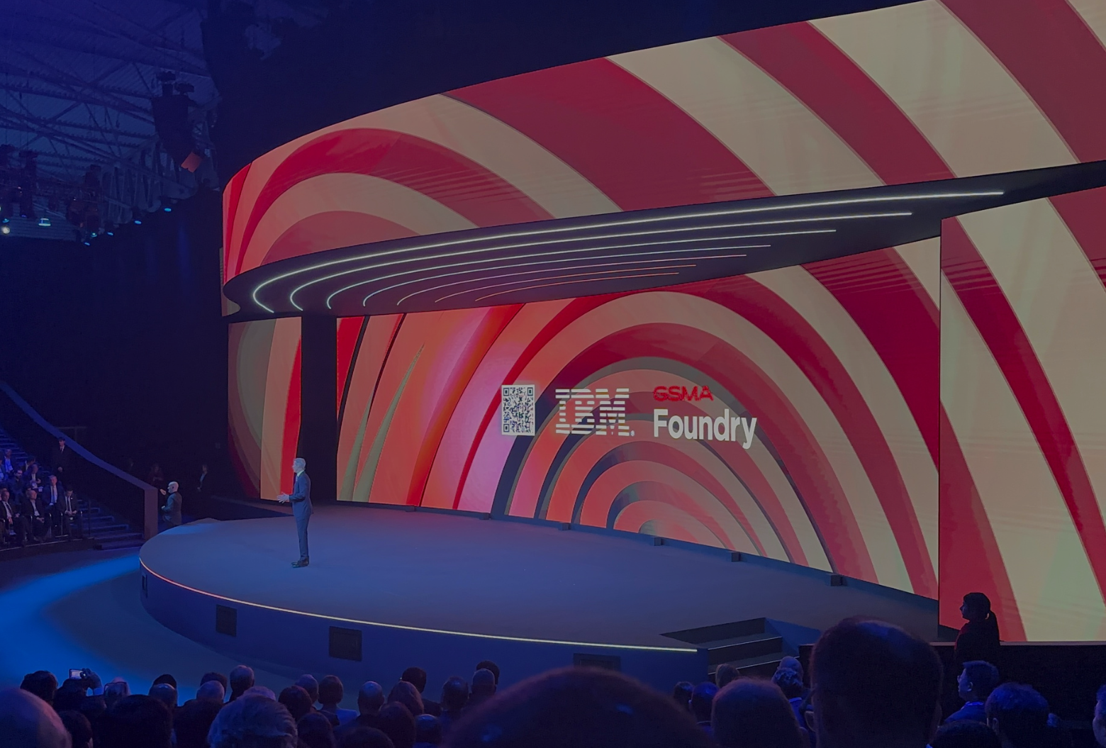 A speaker presents on a curved stage with a large, red and white striped digital screen displaying "gsma foundry" at a tech event.
