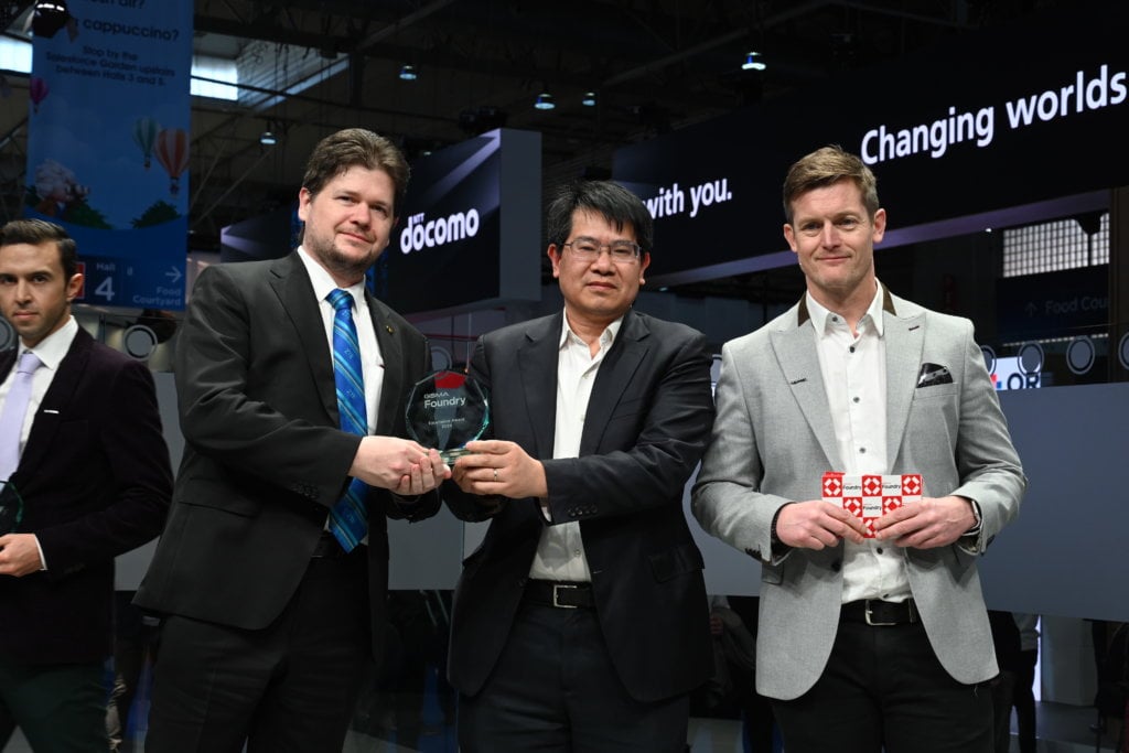 Three men shaking hands and holding an award at a formal event.