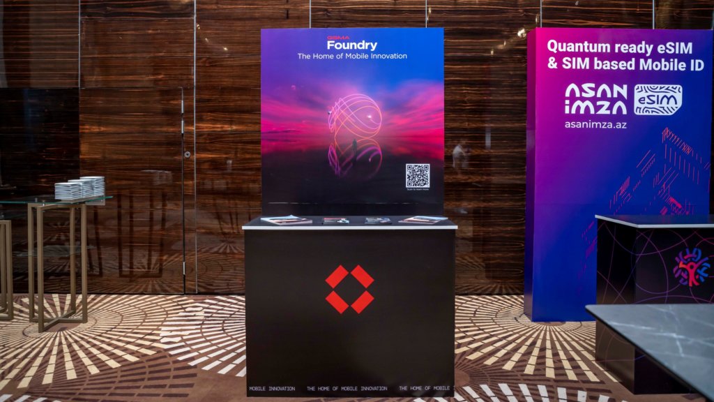 A booth display at a tech event features a standing sign that reads "Foundry - The Home of Mobile Innovation" with a QR code, and "Quantum ready eSIM & SIM based Mobile ID" by Asan Imza. A table in front displays promotional materials.