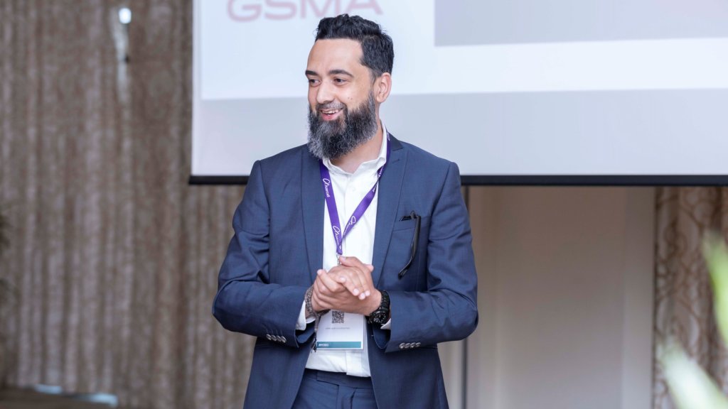 A man in a dark suit and purple lanyard stands in front of a presentation screen with the letters "GSMA" partially visible. He has a beard and is clasping his hands, appearing to speak or present to an audience.