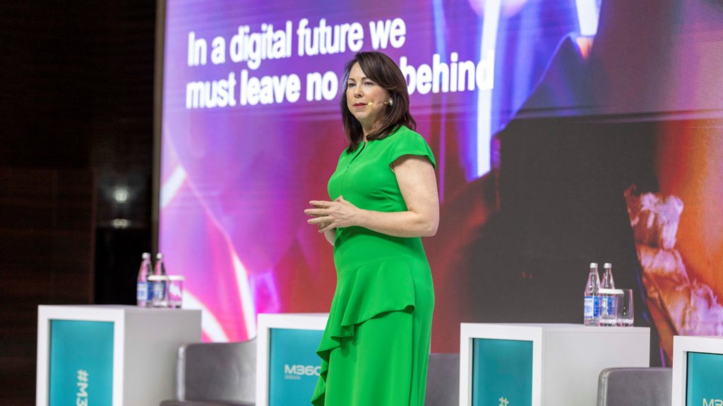 A woman in a green dress stands on a stage, delivering a speech. Behind her is a large screen displaying the text, "In a digital future we must leave no one behind." Bottles of water are placed on white stands next to black chairs on the stage.