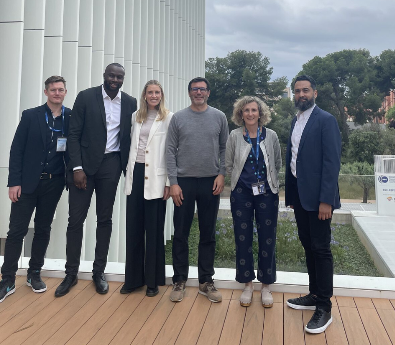 Six people are standing on a wooden deck in front of a modern building. Trees and a cloudy sky are in the background. They are dressed in a mix of business and casual attire, and some are wearing lanyards.
