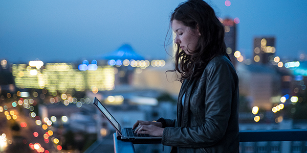 A person with long hair stands on a rooftop at dusk, typing on a laptop. They wear a jacket. The cityscape behind them features blurred lights and buildings under a darkening blue sky, with a glowing pyramid-like structure in the distance.