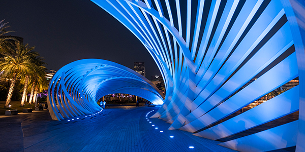 Futuristic blue-lit pedestrian bridge with curved, overlapping metal arches creates a tunnel effect. The pathway is illuminated with small ground lights, with palm trees visible on the left. The night sky is in the background.