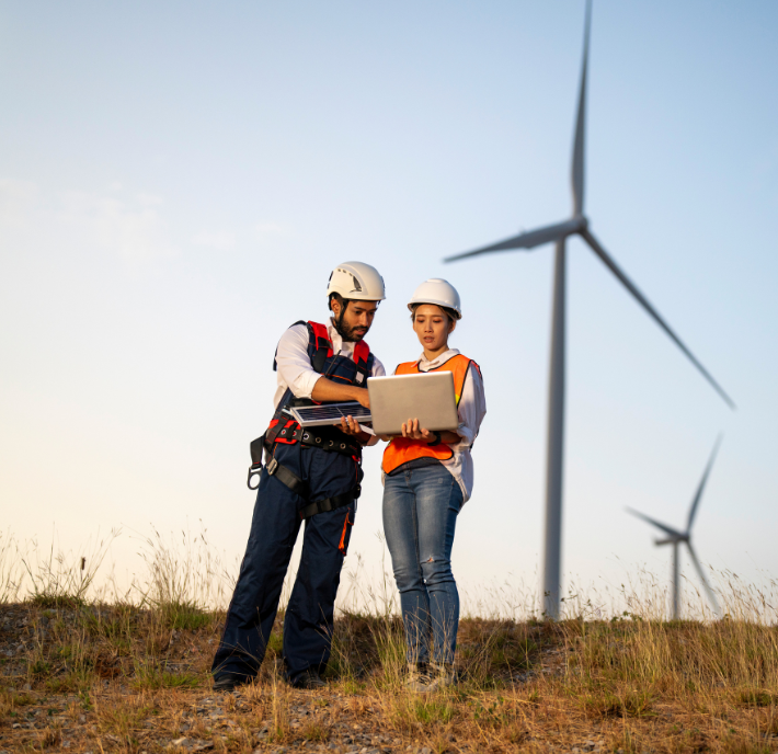 Two engineers wearing safety helmets and vests stand on a grassy hill, discussing a document. A wind turbine is prominently visible in the background, with clear skies above. They appear to be collaborating on a renewable energy project.