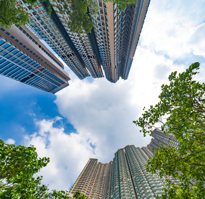 Upward view of tall skyscrapers against a blue sky with white clouds. The buildings, with glass and concrete facades, form a circular frame. Green trees partially surround the bottom edges, adding a contrast between nature and urban architecture.