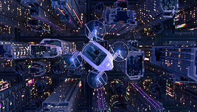 Aerial view of an urban landscape at night with a white drone in the foreground. The drone has four propellers illuminated by blue lights. Below, city streets are lit by streetlights and the windows of tall buildings, creating a vibrant grid pattern.