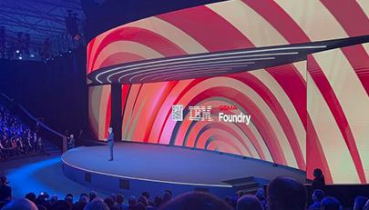 A speaker stands on a curved, elevated stage at a tech event, addressing an audience. Behind, a large screen displays dynamic red and white swirling patterns with the text "GSMA Foundry" and a QR code. The theater is dimly lit, emphasizing the vibrant display.