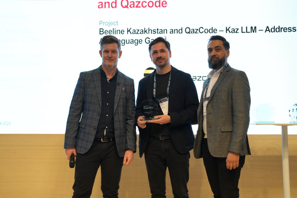 Three individuals on a stage at an awards ceremony. The person in the center holds a glass award, with two others standing on either side. They are dressed in suits. A screen behind them displays text about an award project for Beeline Kazakhstan and QazCode.