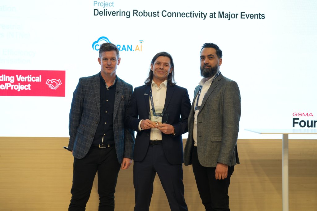 Three men in business attire stand on stage with one man in the center holding a trophy. A presentation slide behind them reads "Project Delivering Robust Connectivity at Major Events." Logos for 5G RAN.AI and GSMA appear beside them. They are smiling, suggesting a positive occasion.