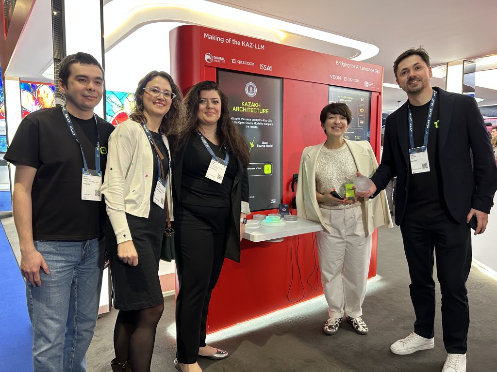 Five people stand smiling in front of a red exhibit display about KAZ-LLaMA at a tech event. Two women hold glass awards. They wear smart-casual attire with badges. The display features text and logos, and the background shows other colorful booths.