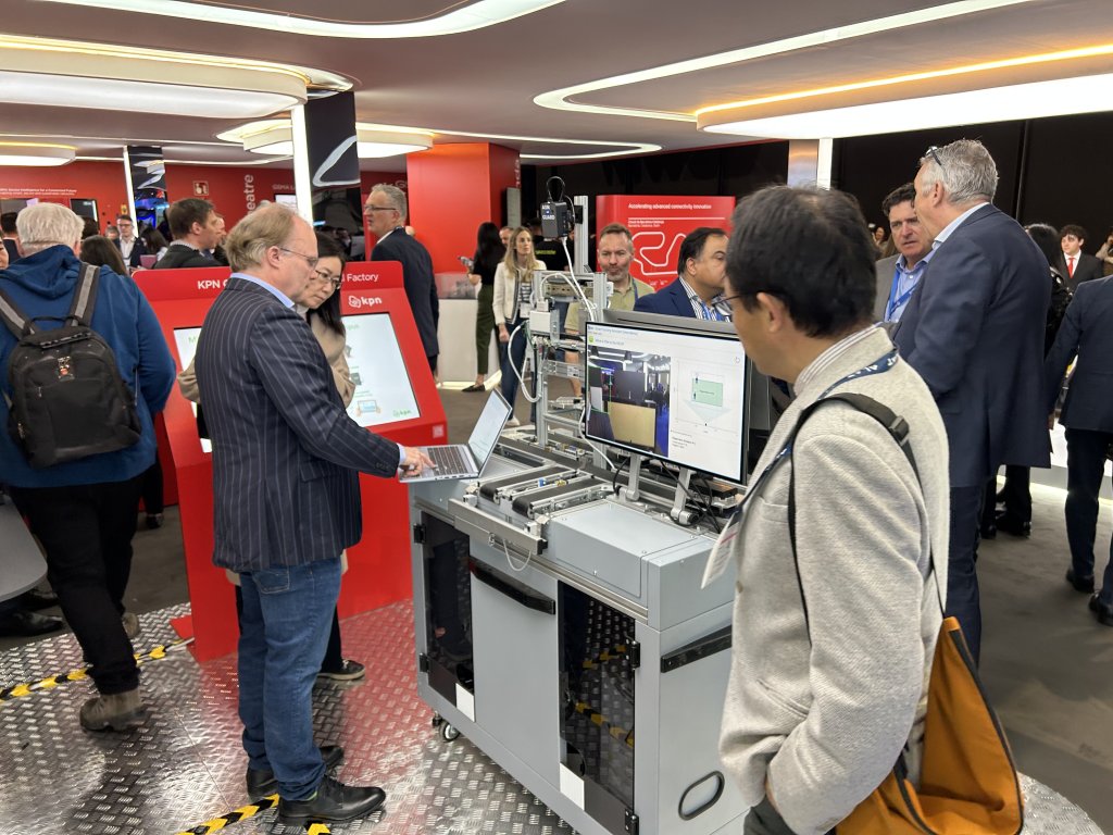 A group of people gather around a high-tech industrial machine at a trade show. Two men in the foreground interact with the device and a laptop, while others converse in the background near bright red and white display booths.