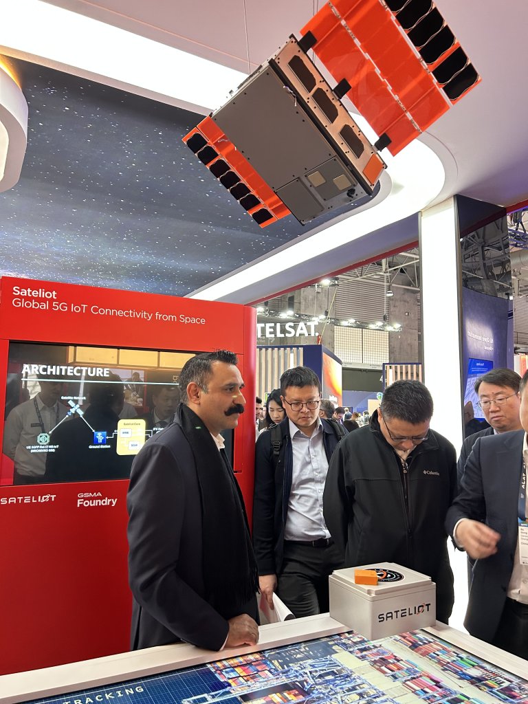 Four men stand around a display booth for Sateliot, featuring a satellite model suspended above. The booth has red branding, technical diagrams, and a sign reading “Global 5G IoT Connectivity from Space.” Other exhibition booths are visible in the background.