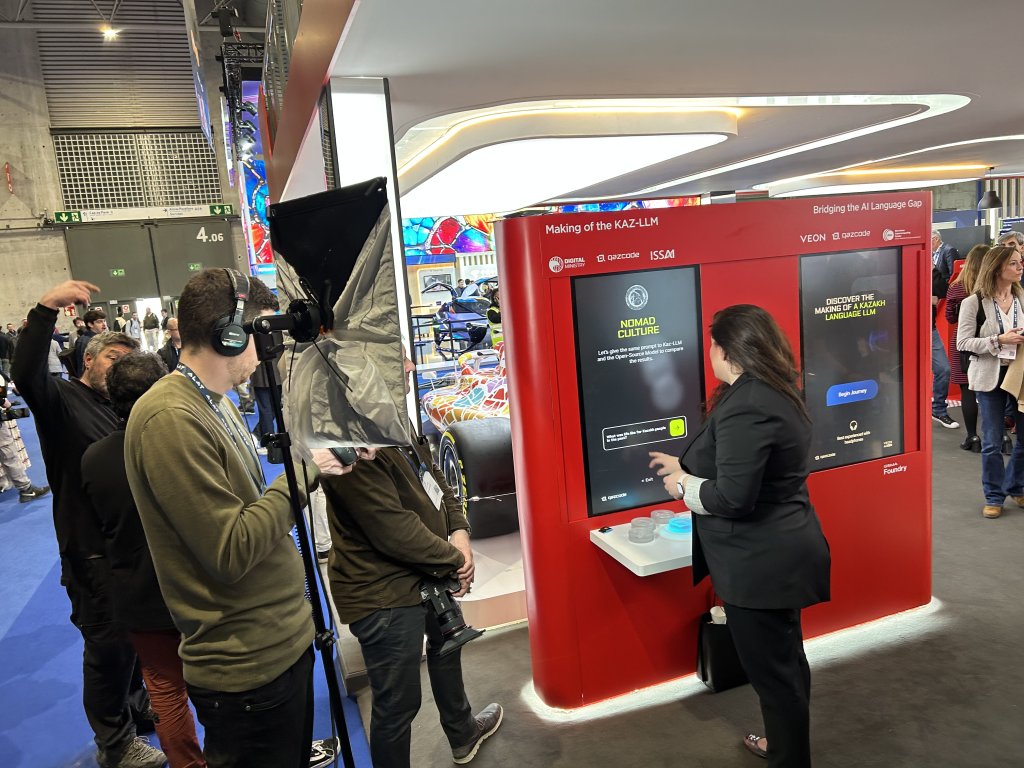 A woman in business attire interacts with a large red digital display booth. A crew with cameras and microphones films her. Bright lighting, a race car, and several people are visible in the background at this busy indoor event or exhibition.