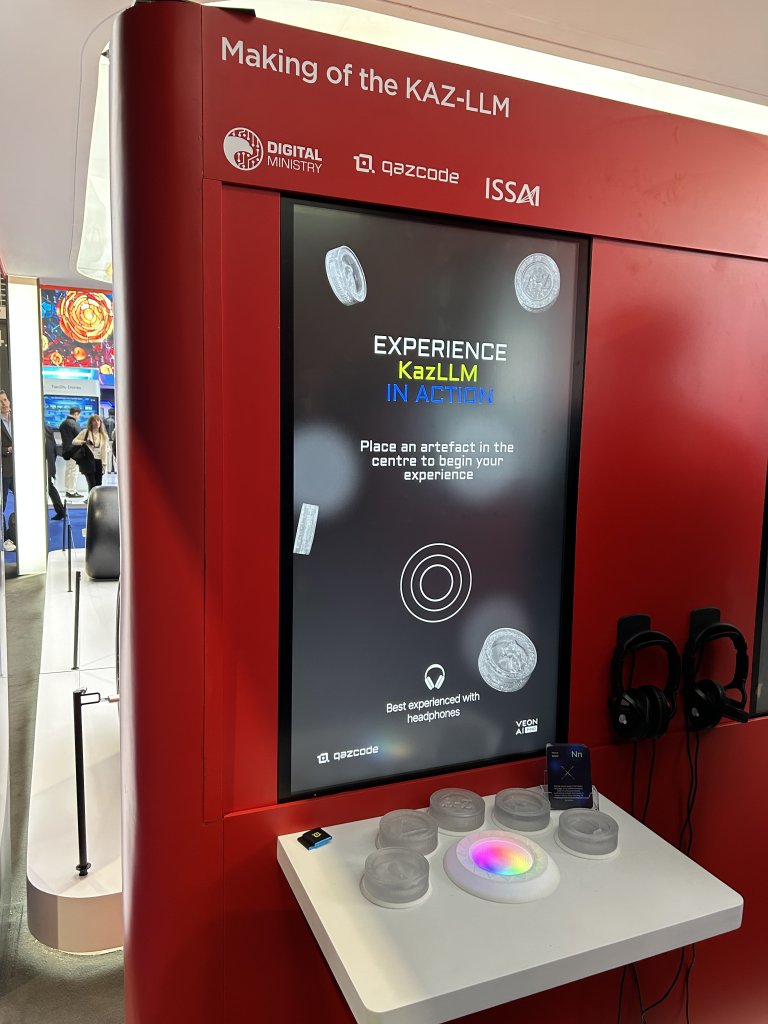 A red kiosk with a large vertical screen reading “Making of the KAZ-LLM." The display invites users to place artifacts in the center to begin an experience. Several round objects and headphones are on a ledge below the screen. Exhibition lights are visible in the background.