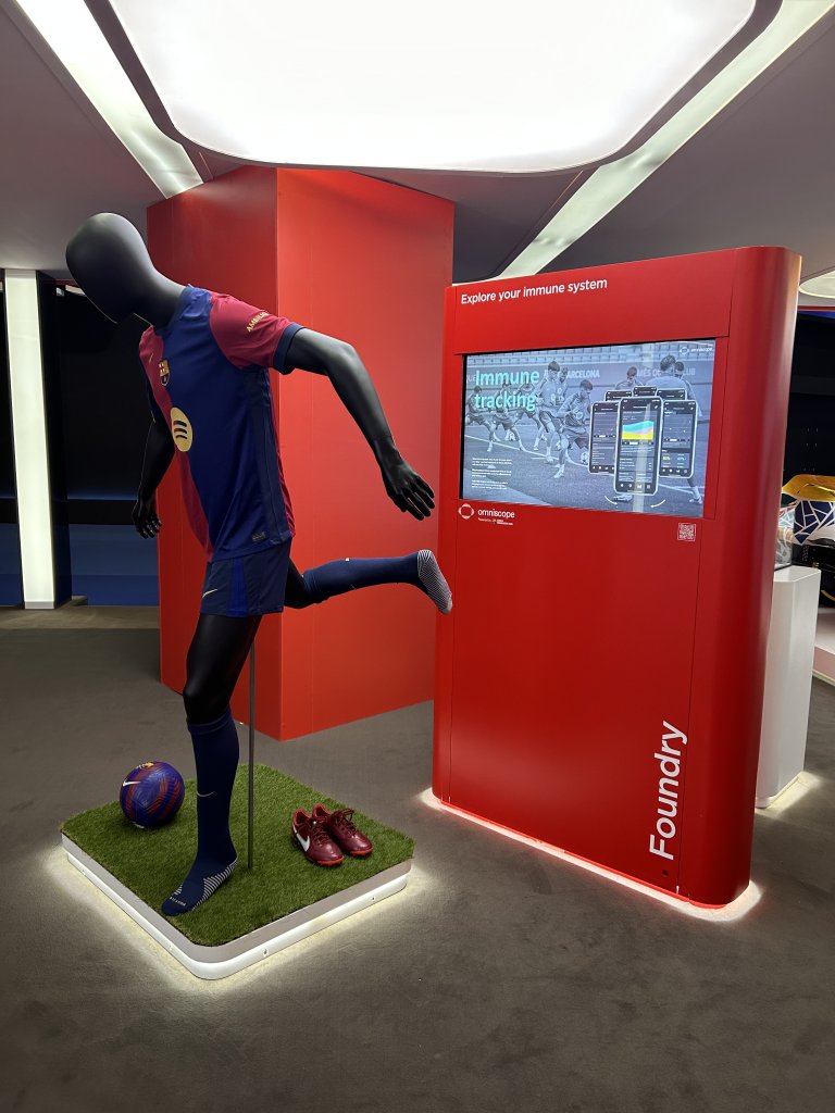 A black mannequin in a red and blue soccer uniform posed as if kicking a ball stands on fake grass near red shoes. Next to it is a red display with a screen showing "Explore your immune system" and digital graphics, in a modern exhibit space.