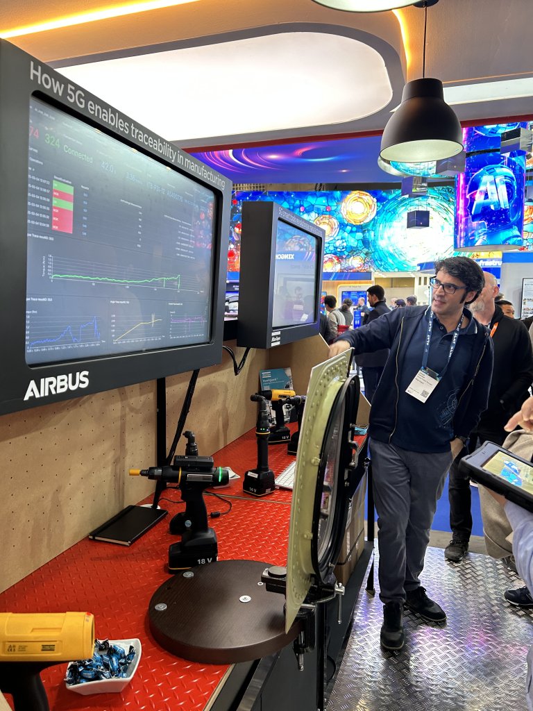 A man in glasses and a badge points at a large screen displaying charts and data about 5G in manufacturing at an Airbus booth. Industrial tools are displayed on a red counter, and bright blue digital artwork decorates the background wall.