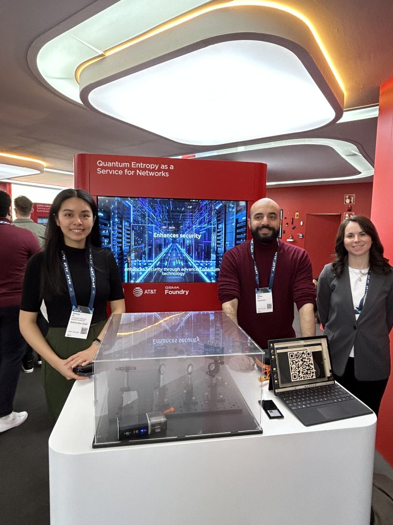 Three people stand smiling at a tech conference booth. Behind them is a screen displaying a data center image and the text "Enhance security." A laptop with a QR code and a transparent device case are on the counter. The booth reads "Quantum Entropy as a Service for Networks.