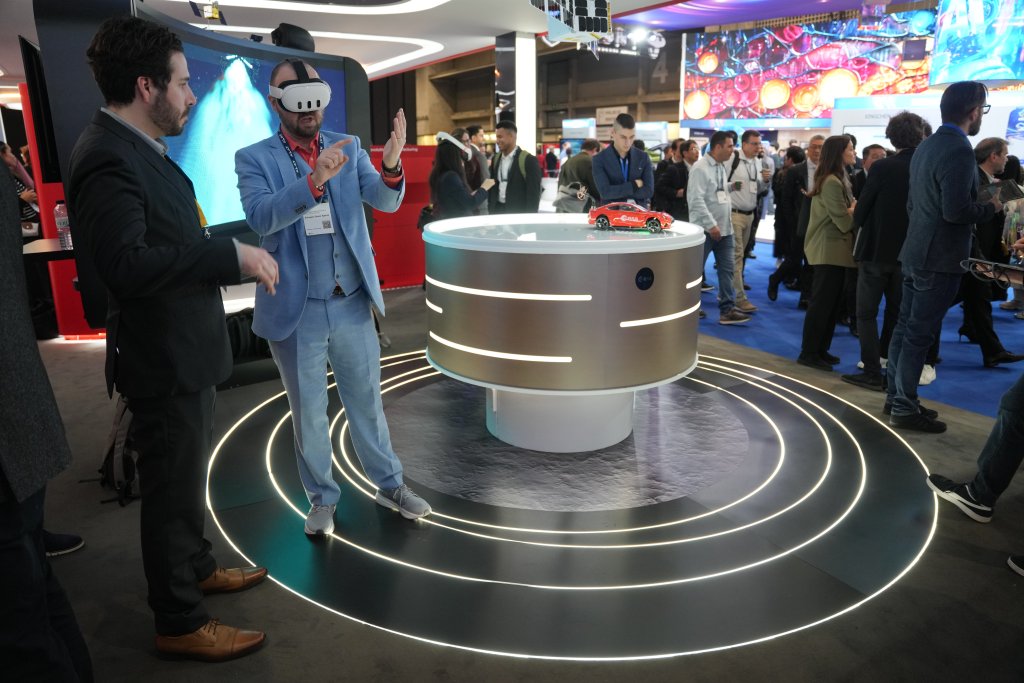 A man in a blue suit wearing a VR headset gestures with his hands while another man watches. They stand near a round display table with a red model car on top, surrounded by glowing circles. People walk and observe technology exhibits in a busy expo hall.