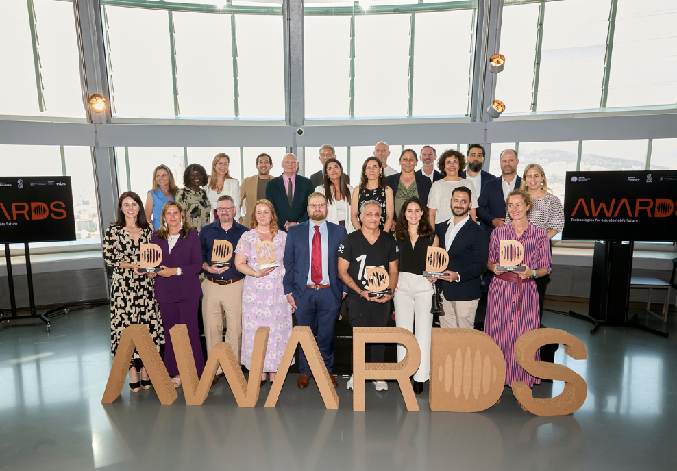 A large group of people in formal attire pose indoors behind giant “AWARDS” letters. Many hold trophy plaques, smiling at the camera. Two screens in the background display the word “AWARDS”. Sunlight streams through tall curved windows behind them.