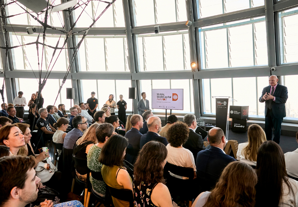 A large group of people sits facing a speaker at a podium in a modern, sunlit room with tall windows. A screen displays “Mobile World Capital Barcelona.” The audience listens attentively, and several people stand near the walls observing the event.
