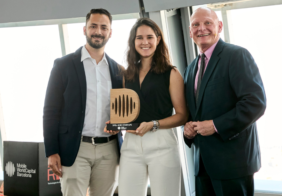 Three people stand indoors, smiling at the camera. The woman in the center holds a wooden award. The two men stand on either side of her, both in suits. Behind them are large windows and a black sign that reads "Mobile World Capital Barcelona.