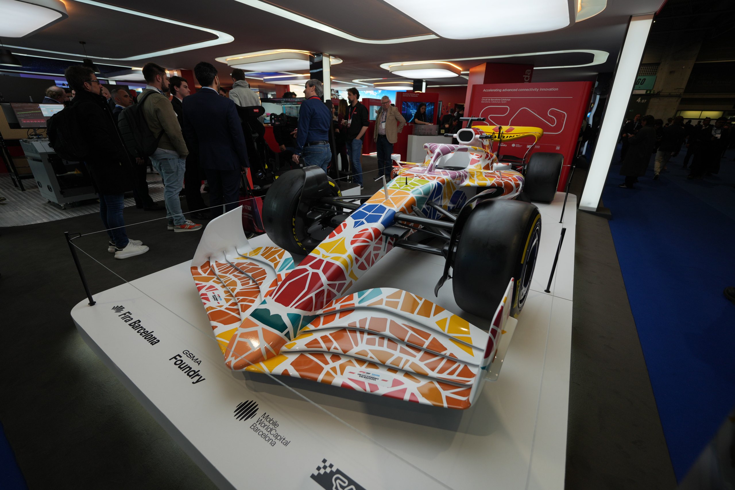 A Formula 1 car with a colorful, geometric mosaic pattern is displayed indoors on a white platform. People stand around the car, which is showcased under modern lighting, and informational signs are visible in the background.