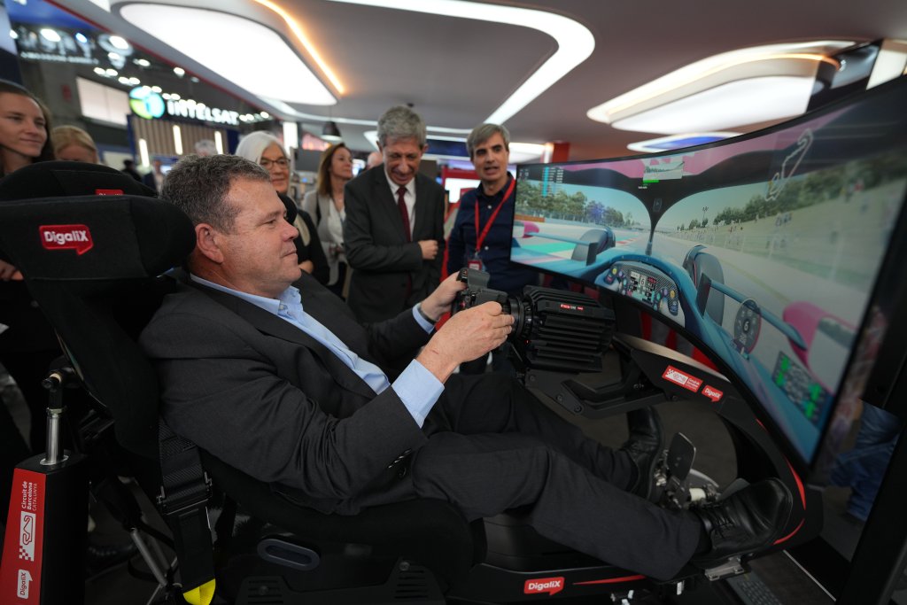 A man in a suit sits in a racing simulator chair holding a steering wheel and pedals, focused on a curved screen showing a driving game. Several people, both men and women, stand around watching. The setting appears to be a tech or gaming expo.