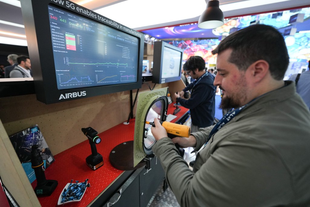 A man uses a yellow power tool on a circular metal component at a display booth. Behind him, a large screen with graphs and data reads “AIRBUS” and shows 5G traceability info. Another person works at a similar station nearby, with people and booths in the background.